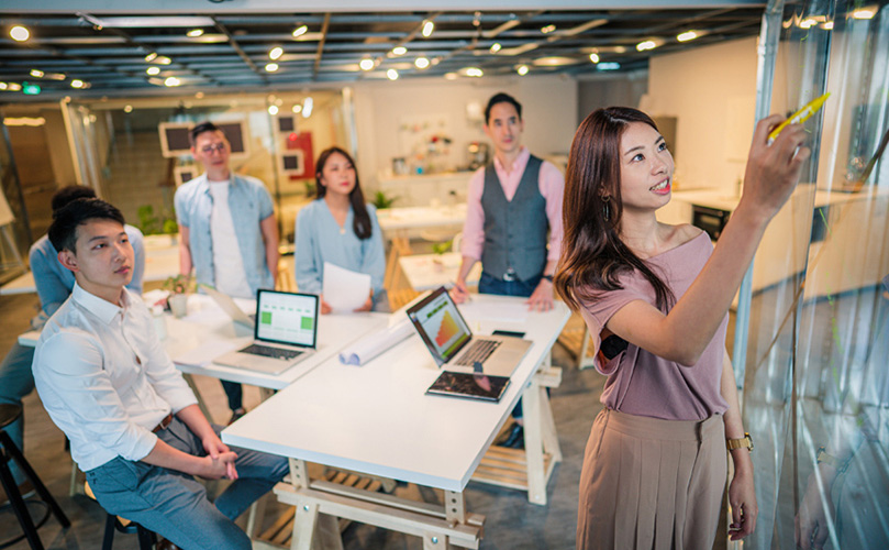 A woman presents using a marker on a glass board to four colleagues in a brightly lit, modern office