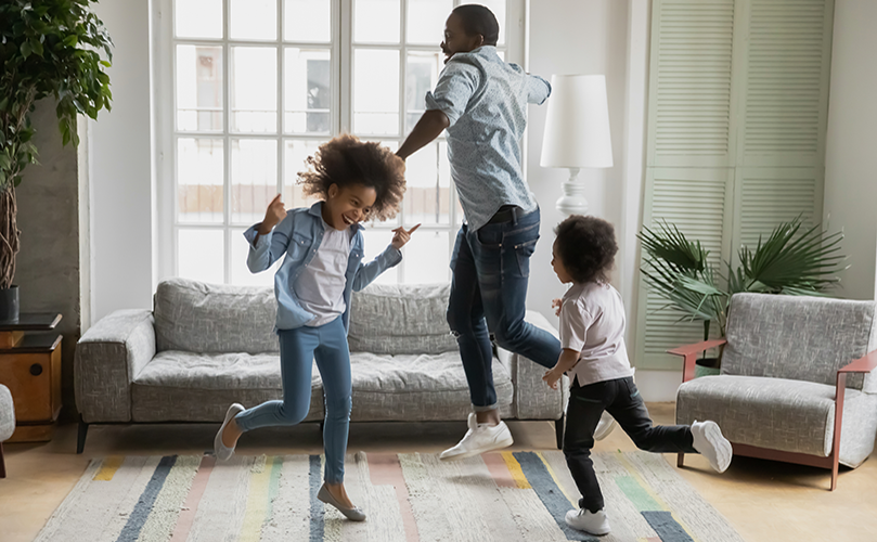 A person and two children are joyfully dancing in a living room with a sofa, armchairs, and plants, in front of large windows.