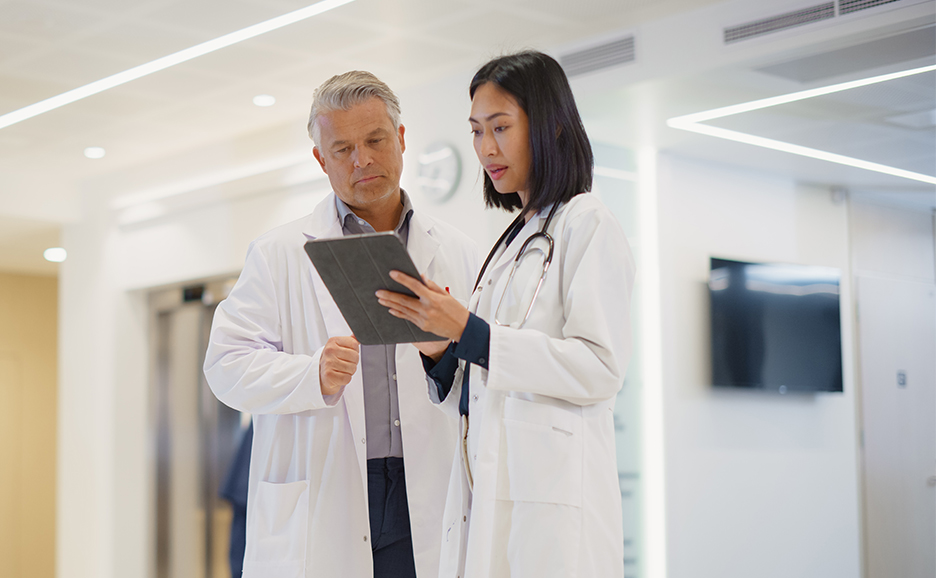 Two doctors in lab coats are discussing while looking at a tablet in a hospital hallway.