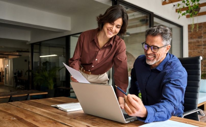 A man and woman collaborate on a laptop in a modern office setting, focused on their work.