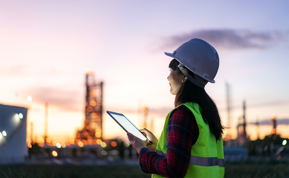 ヘルメットと安全ベストを着用した女性が、夕暮れの工場を背景にタブレットを操作している様子