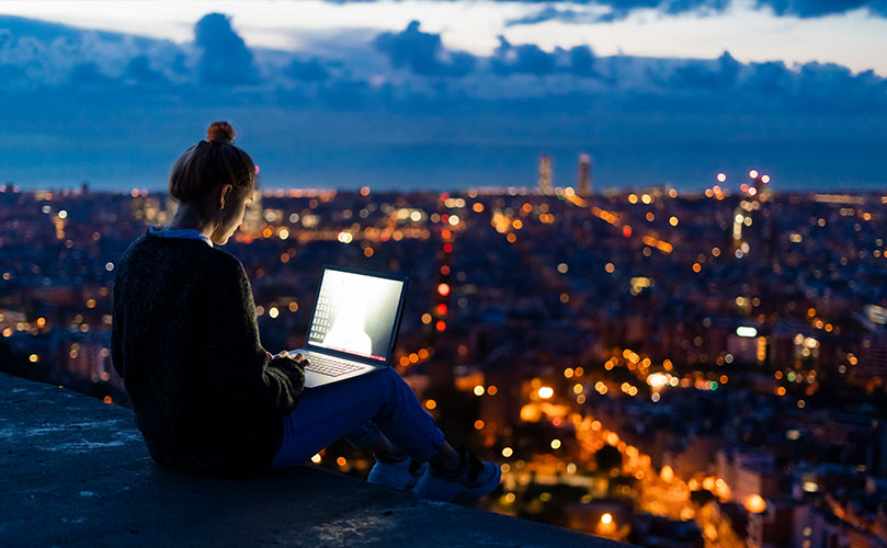 A woman sits on a ledge at night, focused on her laptop, illuminated by the screen's glow against the dark backdrop.