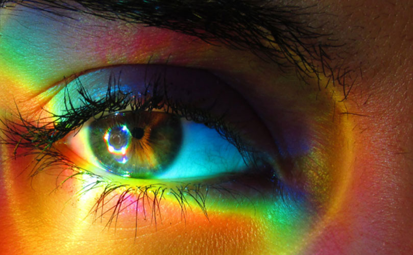 Close-up of a human eye with rainbow colours reflected on the skin and eyelashes.