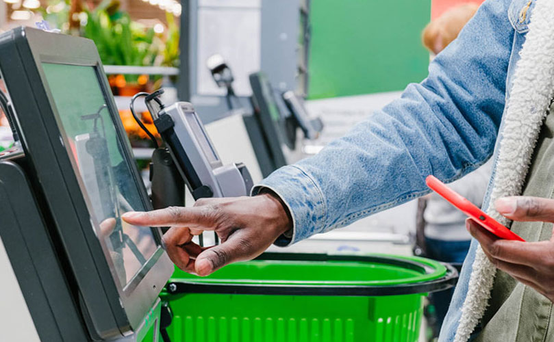 Close-up of a person in a denim jacket using a self-service checkout touchscreen in a supermarket, with a green shopping basket beside them.