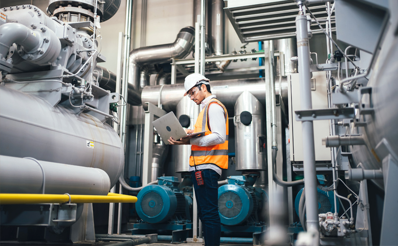 Engineer in a hard hat and reflective vest using a laptop in an industrial plant with large machinery and pipelines around