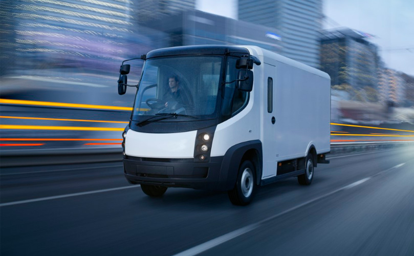 A white van travels down a road with blurred city buildings in the background.