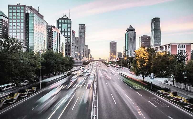 A cityscape with a motorway running through the centre, flanked by tall buildings. Blurred images of cars represent movement and speed.