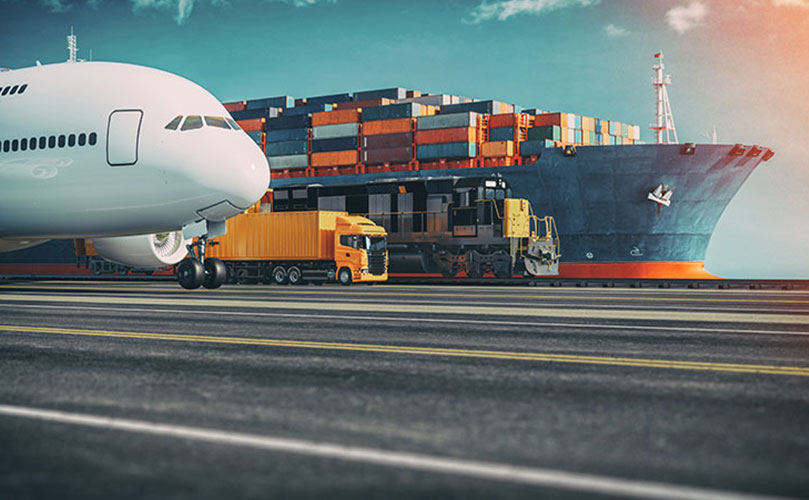 In the distance a large cargo ship is being loaded with containers, while a train and yellow truck sit alongside it. In the foreground, there is the nose of an airliner.