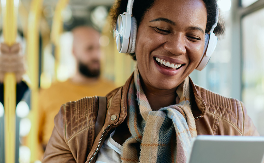 A woman sitting on a bus with headphones, smiling while looking at a tablet she's holding