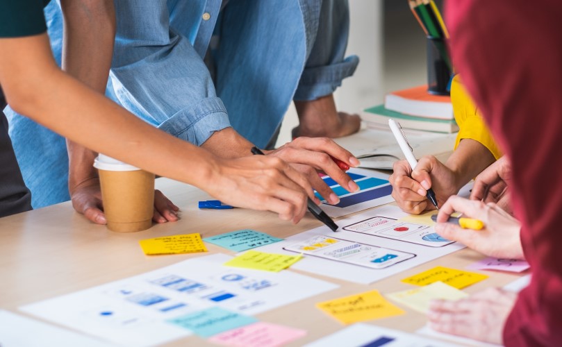 A desk full of papers and post its with peoples' hands writing on them