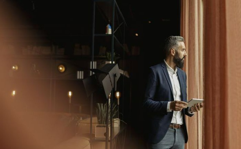 A suited man gazes at his phone while standing next to a window.