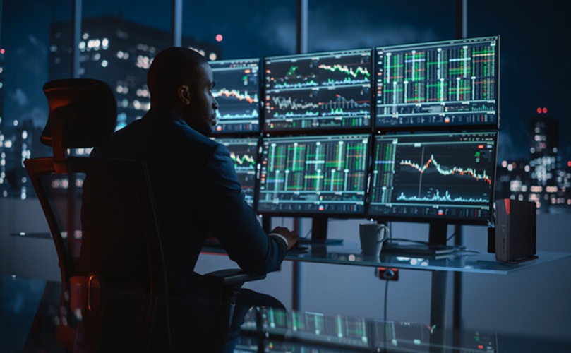 A man seated at a desk with several monitors showing real-time stock market information and analytics.
