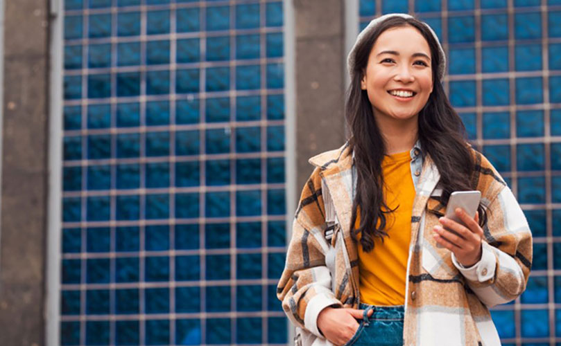 A smiling young woman with long dark hair, wearing a yellow top and patterned jacket, holds a mobile phone in front of a glass-fronted modern building.