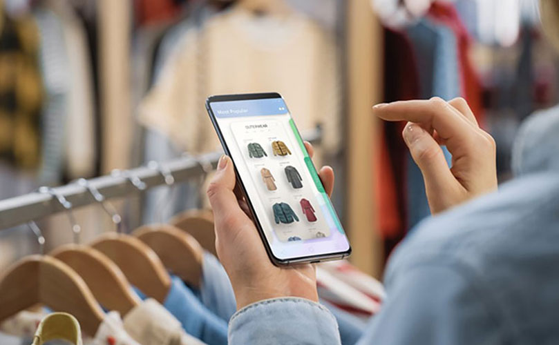 A person in a clothing store browses jackets on a smartphone shopping app, with blurred garments hanging in the background.