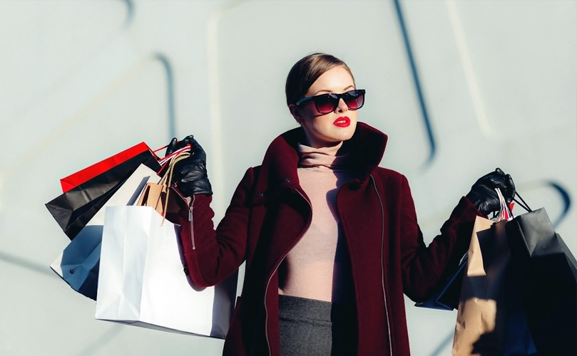 A fashionable woman in a burgundy coat, wearing sunglasses and red lipstick, carrying shopping bags