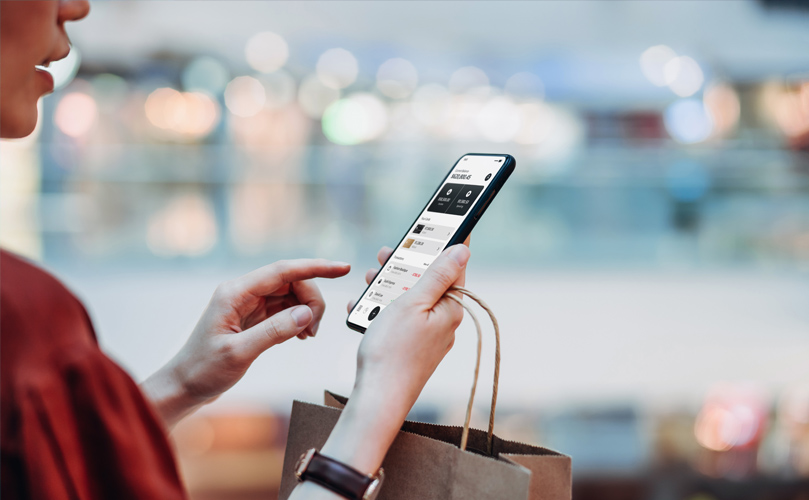 Une personne faisant ses courses avec un smartphone, vu de côté, tenant un sac à provisions dans une main et interagissant avec un smartphone grand écran avec l'autre.