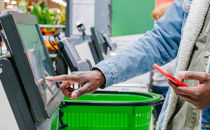 Close-up of a person in a denim jacket using a self-service checkout touchscreen in a supermarket, with a green shopping basket beside them.