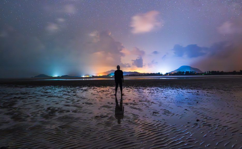 夜の砂浜に立つ人影、遠くに街の灯りが見える風景。