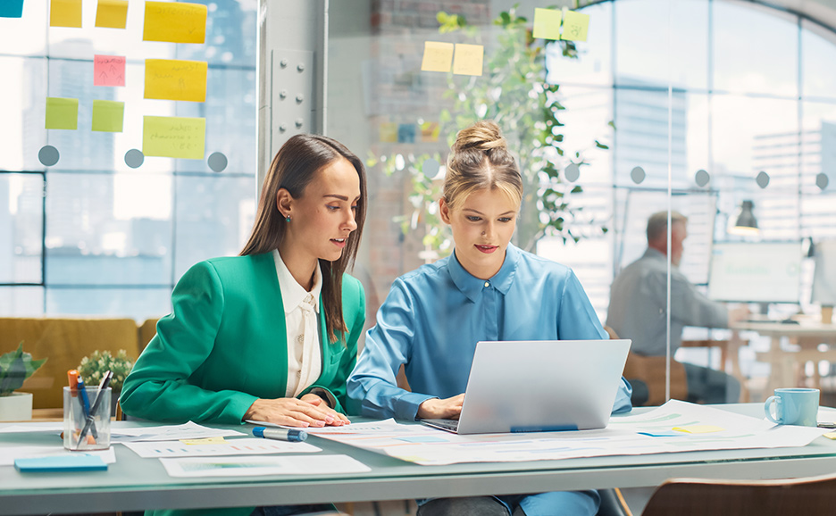 Two women collaborating in a bright office, with one woman in a green jacket pointing at the laptop screen of the woman next to her.