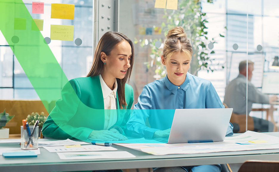 Zwei Frauen arbeiten in einem hellen Büro zusammen, wobei eine Frau in einer grünen Jacke auf den Laptop-Bildschirm der Frau neben ihr zeigt.