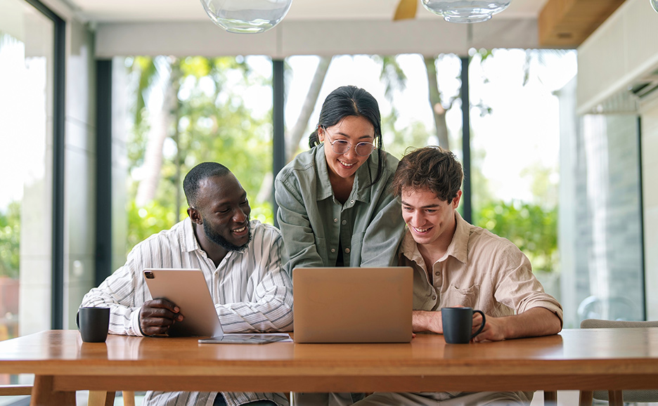Three smiling colleagues sitting at a wooden table, looking at a laptop.