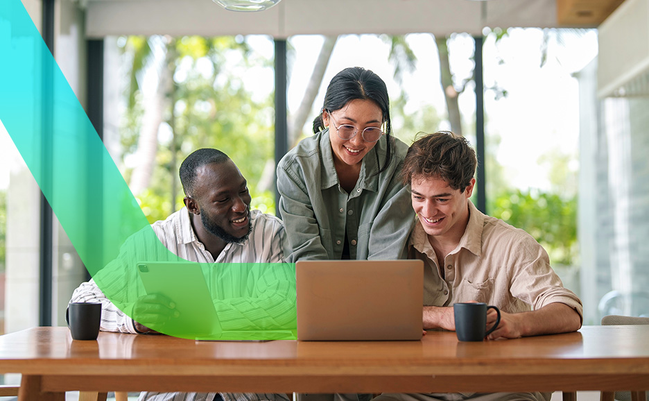 Three smiling colleagues sitting at a wooden table, looking at a laptop.