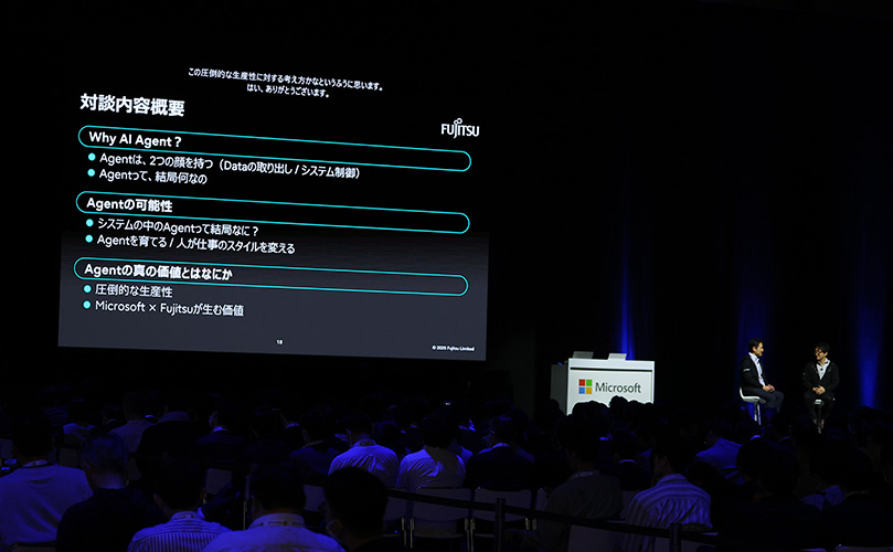 プレゼンテーションのスライドを映し出したスクリーンを観客席から撮影した写真です。スライドには日本語で「対談内容概要」とあり、以下のような箇条書きが記されています。 Why AI Agent? Agentは、2つの顔を持つ(Dataの取り出し / システム制御) Agentって、結局何なの Agentの可能性 システムの中のAgentって結局なに? Agentを育てる / 人が仕事のスタイルを変える Agentの真の価値とはなにか 圧倒的な生産性 Microsoft × Fujitsuが生む価値 壇上にはMicrosoftのロゴがついた演台があり、二人の人物が座って対談している様子が薄暗く映っています。観客席には多くの人が座っており、後姿が見えています。会場は全体的に暗い照明で、壇上は青い照明で照らされています。