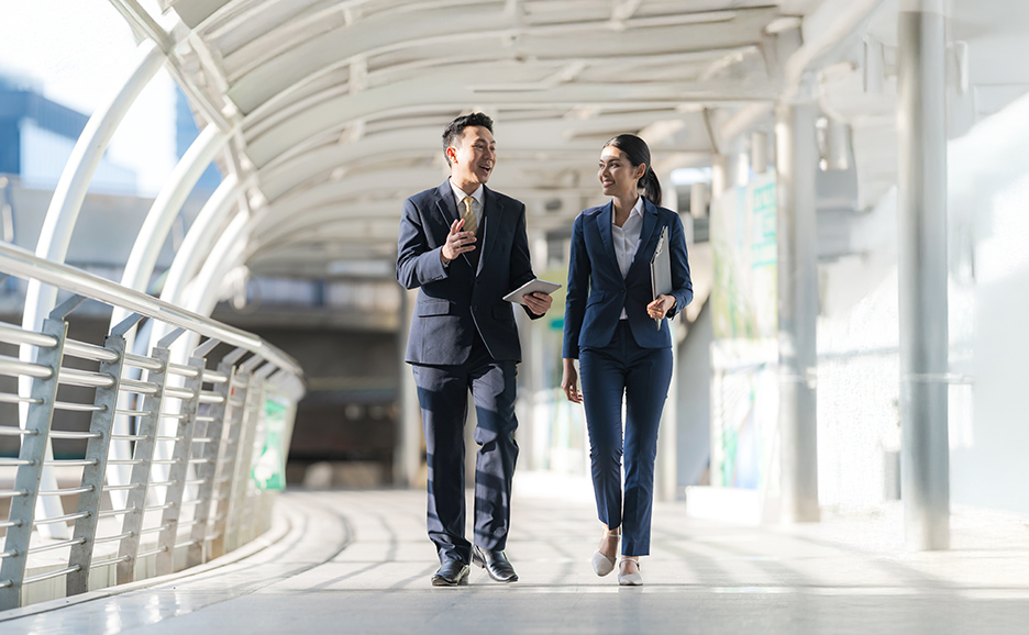 A man and a woman in business suits walk and talk on an outdoor walkway. The man holds a tablet.