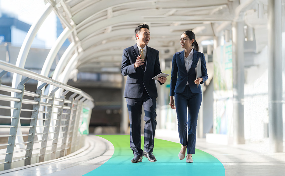 A man and a woman in business suits walk and talk on an outdoor walkway. The man holds a tablet.