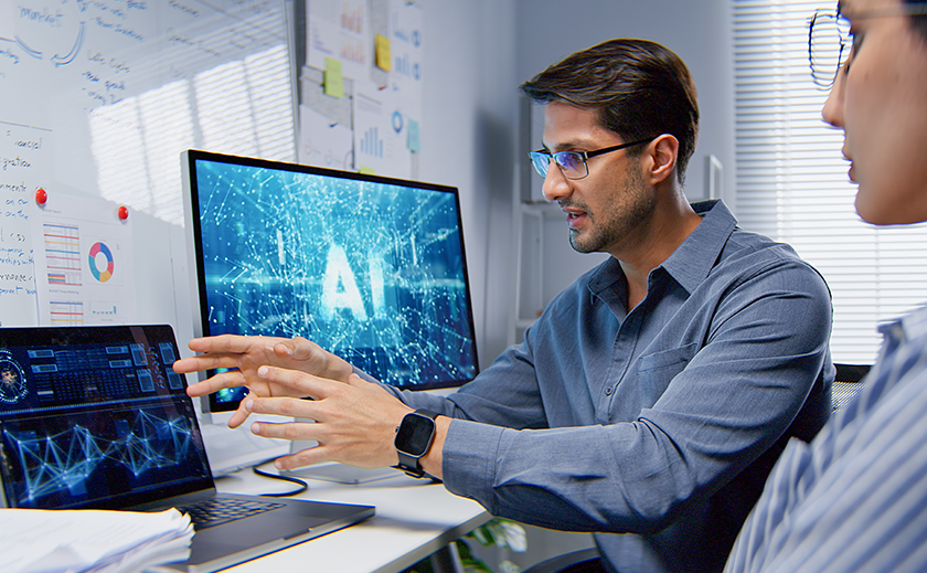 Two professionals discussing data analytics and artificial intelligence (AI) on laptops and a large monitor displaying 'AI' text and network graphics in a modern office."