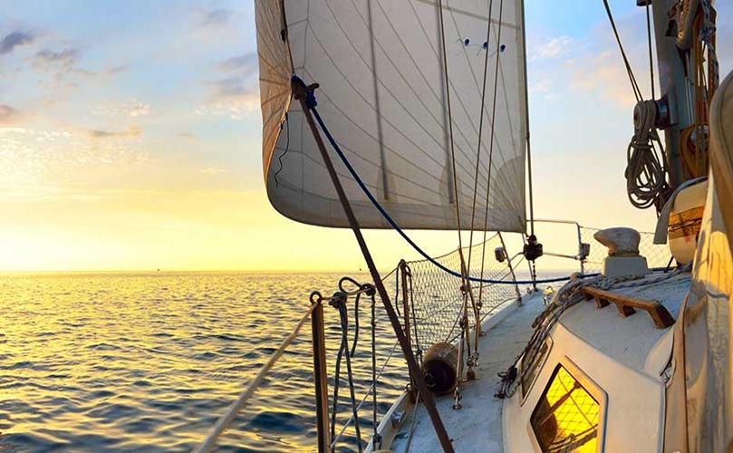 A sailboat navigates the ocean waters as the sun sets, casting warm hues across the sky.