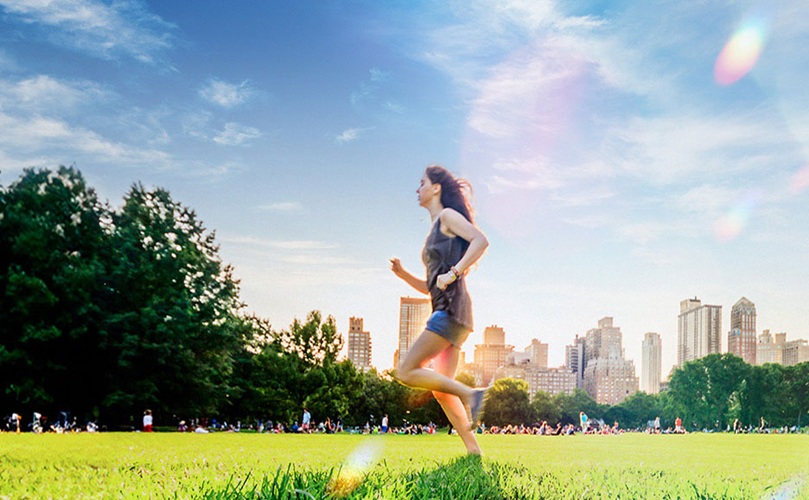 A woman jogs through a park, with a city skyline rising in the background, blending nature and urban scenery.