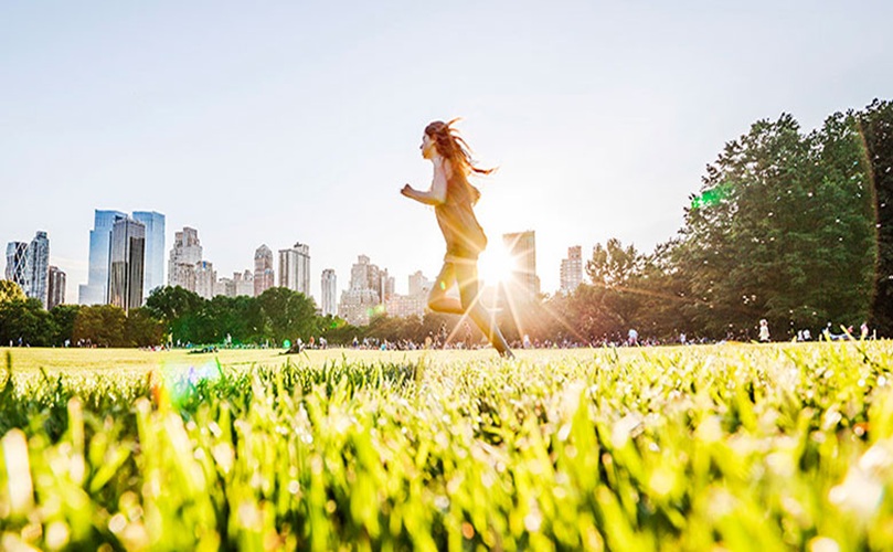 A woman jogs through a park, sunlight shining brightly behind her, illuminating the scene.