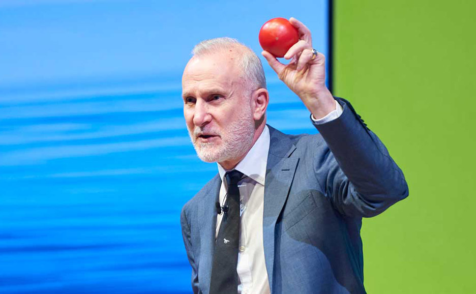 白い髪と髭を生やした、スーツを着た男性（BWT社のCEO、テリー・ポール氏）がトマトを片手に講演を行う様子