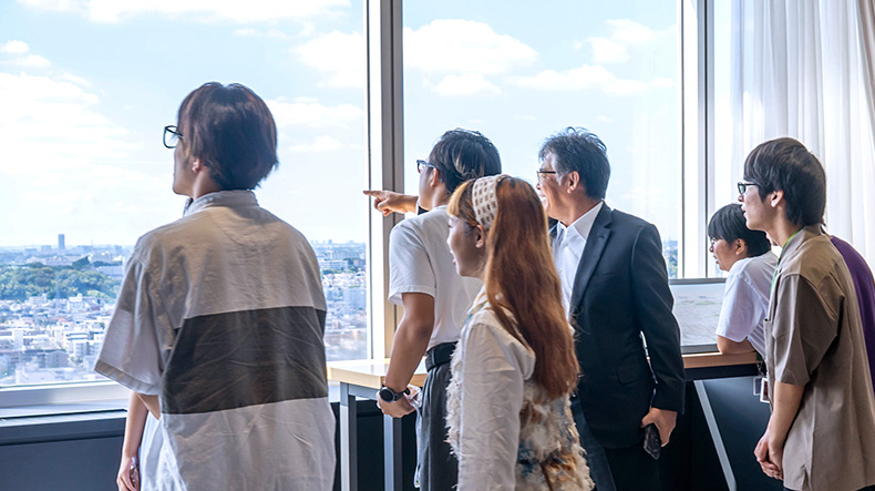 開けた景色が見える窓際で5名の生徒と時田社長が景色を眺めている。