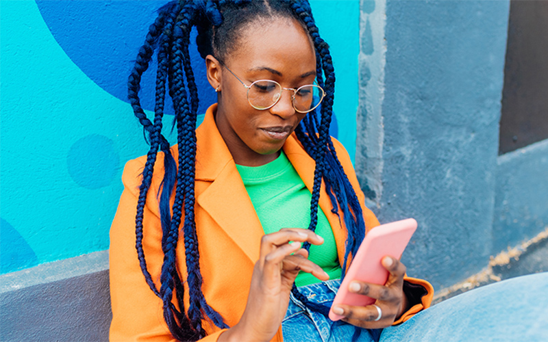 A woman wearing glasses and dreadlocks is focused on her phone screen.