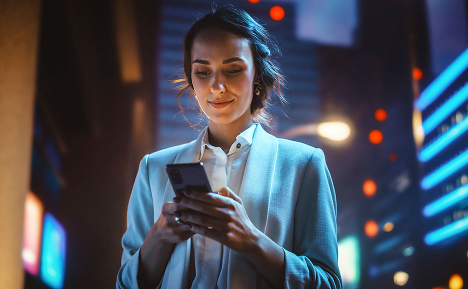 Frau in Businesskleidung nutzt ein Smartphone vor einer beleuchteten Stadtkulisse bei Nacht