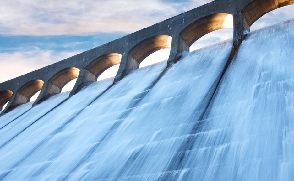 Nahaufnahme einer Staudammkrone mit stark herabfließendem Wasser und bogenförmigen Öffnungen vor blauem Himmel