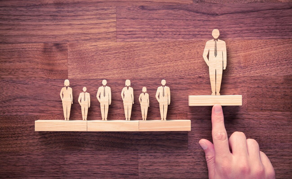 Auf einem Holztisch stehen 6 kleine und eine große Holzfigur von Menschen. Eine Hand zeigt auf die große Holzfigur