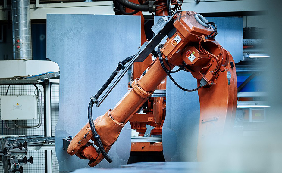Industrieroboterarm in einer Produktionshalle, der ein Metallbauteil automatisiert bearbeitet.