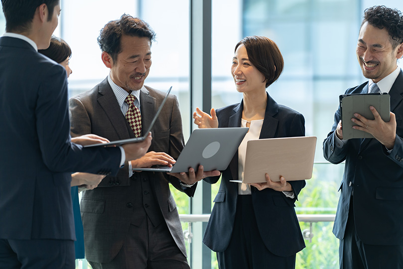 複数のビジネスパーソンが、ノートパソコンやタブレットを手に、明るいオフィスで笑顔で話し合っている様子。