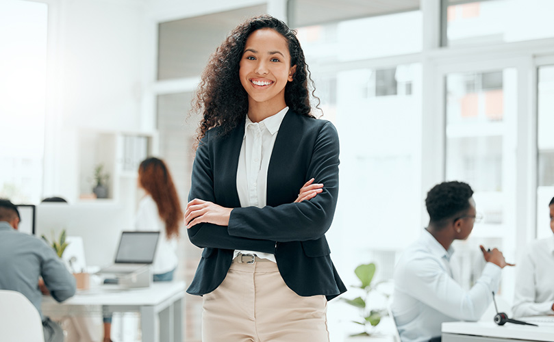 オフィスの混雑した背景を背に、腕を組んで笑顔で立っている女性