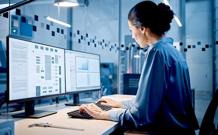 研究室やオフィスのような場所で、女性が2つのモニターを使ってコンピューターで作業している