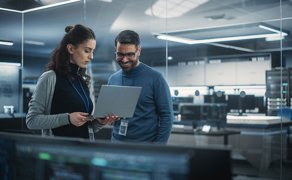 データセンターでノートパソコンを共有する男女。真剣な表情の女性と笑顔の男性。