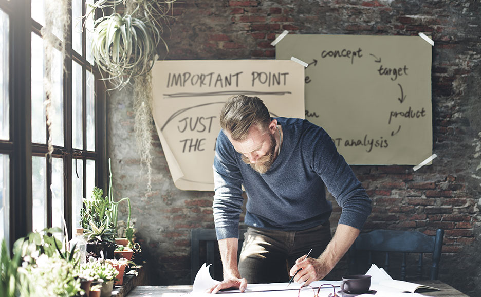 ひげを生やした男性が、プランニングボードの貼られたレンガ壁の前で図面を引いている