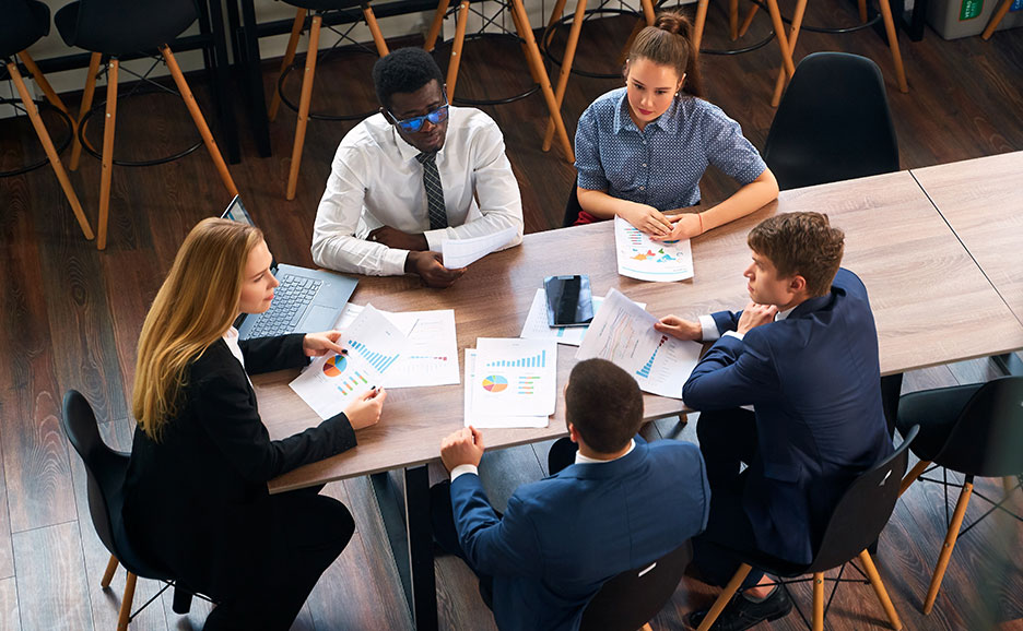 木製のテーブルを囲んで会議をしている5人のビジネスパーソン。