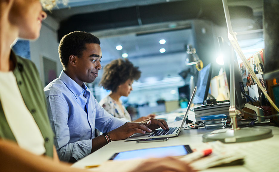 オフィスでパソコンに向かう人々。手前の男性が笑顔でキーボードを打っている。