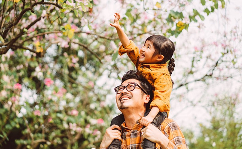 An adult carries a smiling child on their shoulders outdoors, The child reaches up toward tree branches with pink flowers.