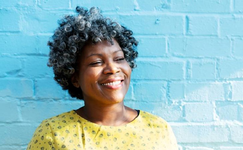 A smiling woman with curly hair stands against a blue wall, radiating joy.