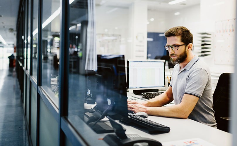 A man in glasses is diligently working on a computer, looking at the screen and typing with intent.
